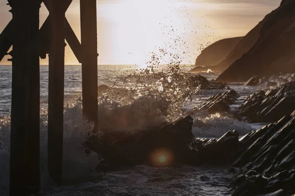 日落时分，在木码头下的海岸上，美丽的海浪在尖尖的岩石上 — 图库照片