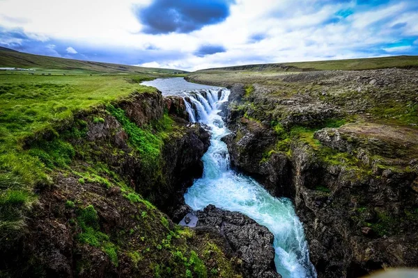 Hermosa toma de cascada que fluye hacia abajo en medio de colinas rocosas bajo un cielo nublado — Foto de Stock