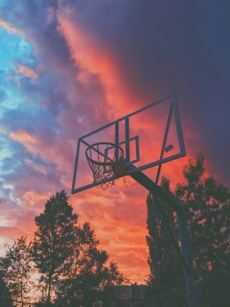 Tiro vertical de un aro de baloncesto en un campo con un hermoso cielo rojo en el fondo al amanecer — Foto de Stock