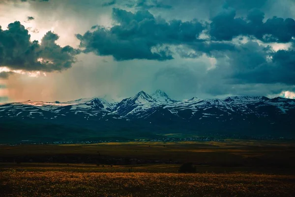 Szerokie ujęcie pięknego pola z zapierającymi dech w piersiach pokrytymi śniegiem skalistymi górami z niesamowitymi chmurami — Zdjęcie stockowe