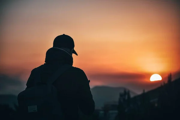 Detrás de la toma de un misterioso macho en una gorra de béisbol y una mochila mirando el hermoso atardecer —  Fotos de Stock