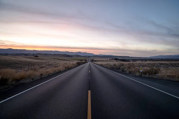 El disparo de una carretera rodeada de campos de hierba seca bajo el cielo durante la puesta del sol —  Fotos de Stock