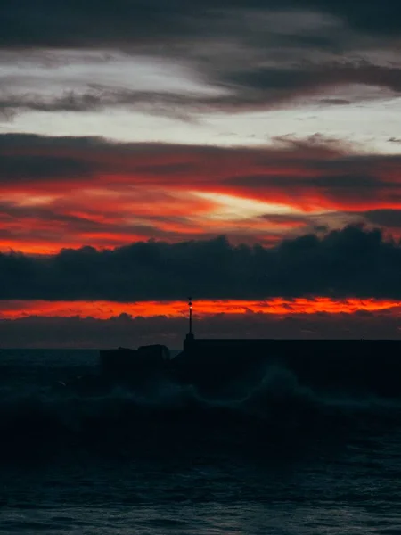 Tiro vertical de uma onda do mar com uma silhueta de edifício na distância e céu vermelho no pôr do sol — Fotografia de Stock