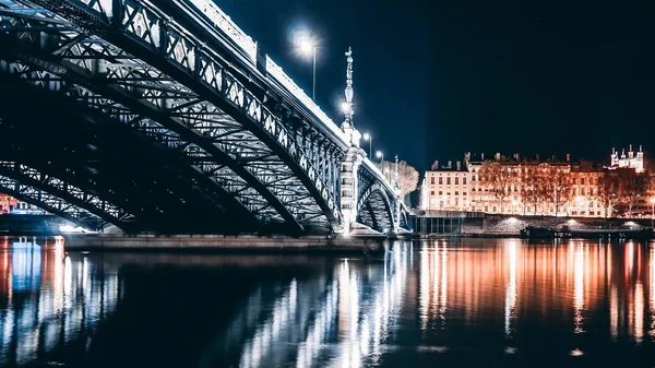 Hermosa toma de un largo puente de acero sobre un río con luces y reflejos en el río por la noche —  Fotos de Stock