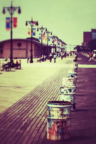 Vertical shot of people walking on a pathway near metal barrels and lamp posts — Stock Photo, Image