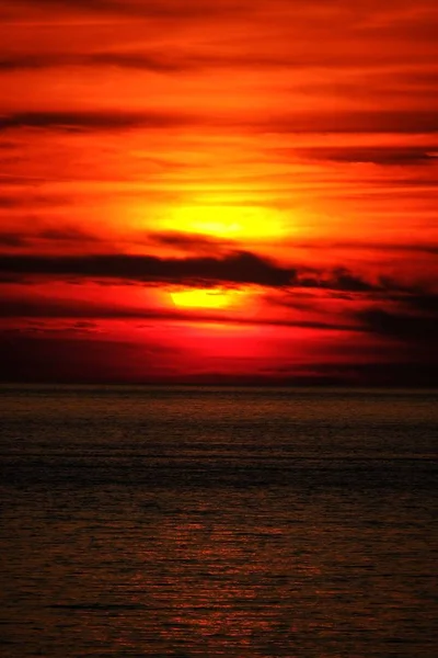 Vertical shot of a sea and sun behind the clouds with a red sky at sunset — Stock Photo, Image