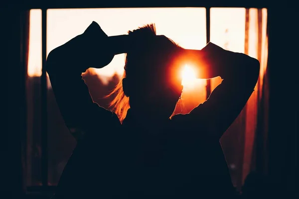 Silhouette de una mujer que fija su cabello delante de una ventana con puesta de sol en el fondo. — Foto de Stock