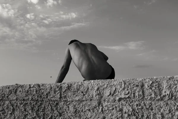Полуобнаженный человек, сидящий на камне на пляже спиной к камере — стоковое фото