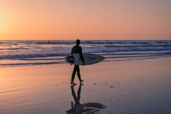 Maschio in possesso di una tavola da surf a piedi sulla spiaggia al bellissimo tramonto al mare — Foto Stock