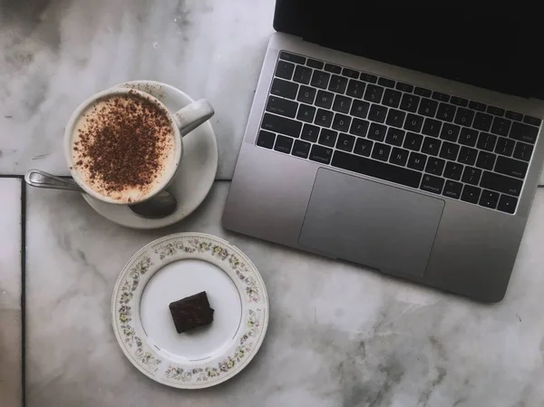 Overhead shot of a laptop with a cappuccino and a brownie on a ceramic surface