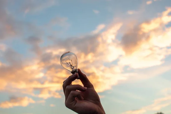 Personens hand håller upp en liten glödlampa på en vacker vit molnig himmel i bakgrunden — Stockfoto