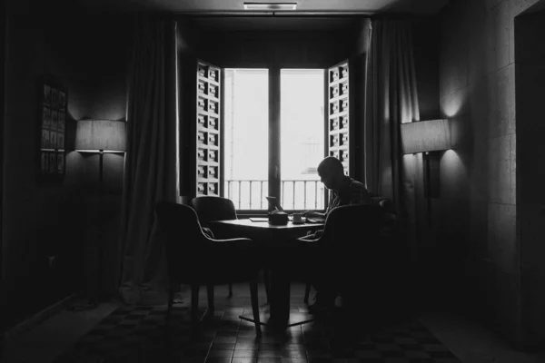 一个男人坐在靠近敞开窗户的桌子旁边的椅子上的黑白照片 — 图库照片