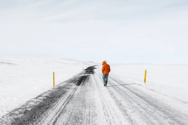 Breed beeld van een man in een oranje jasje die over een besneeuwde snelweg loopt — Stockfoto