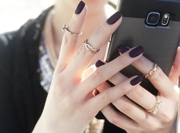 Female with black nail polish and chain-like rings holding a smartphone.