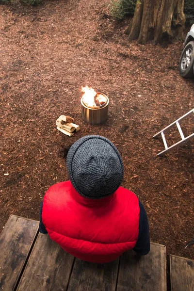 Una persona con sombrero gris y chaleco rojo aislado sentada junto a una leña ardiendo en una olla — Foto de Stock