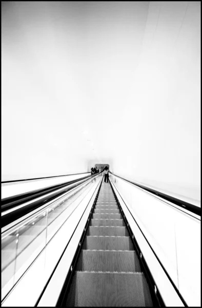 Scatto verticale di una scala mobile che scende con persone in piedi in lontananza in bianco e nero — Foto Stock