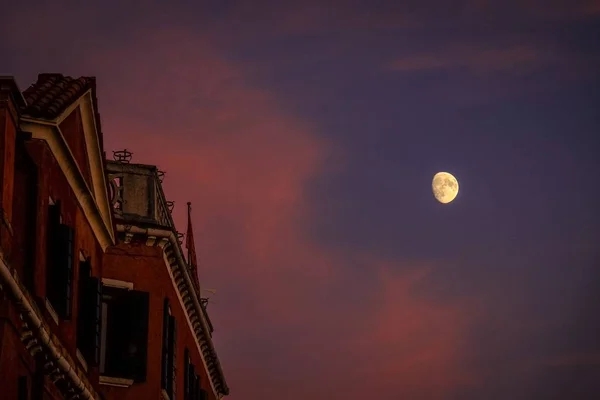 Tiro de ângulo baixo de um edifício alto com uma lua visível no céu roxo e azul — Fotografia de Stock