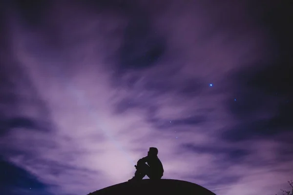 Belo tiro largo de uma pessoa sentada em uma rocha sob um céu estrelado noite púrpura — Fotografia de Stock