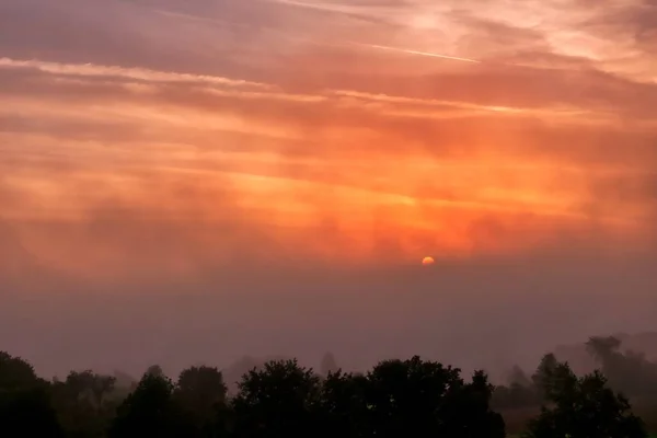 Lindo tiro do pôr do sol incrível com o céu vermelho sobre uma floresta enevoada no campo — Fotografia de Stock