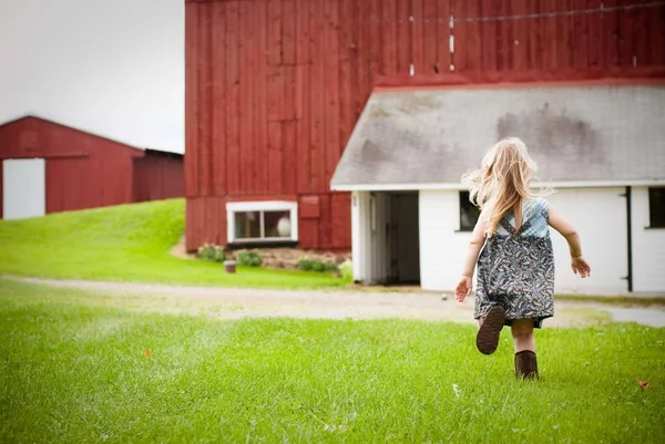 Beautiful shot of young girl running on the grass towards a large red wooden barn — Stock Photo, Image