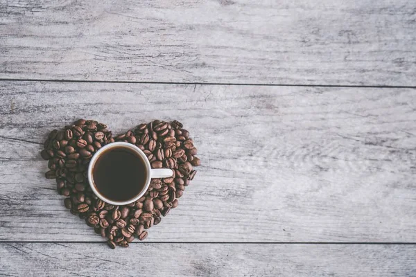 Fotografia aérea de grãos de café em forma de coração com uma caneca de café em um fundo de madeira — Fotografia de Stock