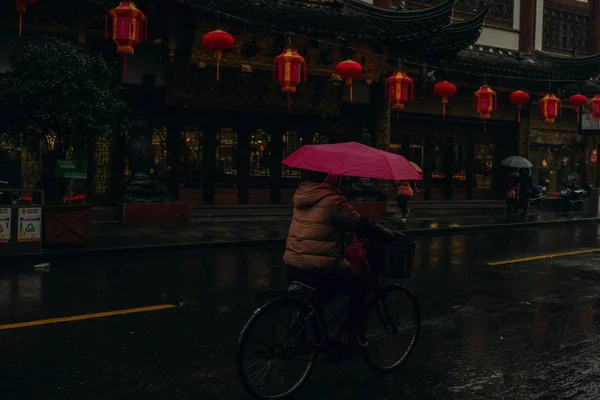 Pessoa segurando um guarda-chuva rosa andando de bicicleta em uma rua molhada perto de um edifício tradicional chinês — Fotografia de Stock