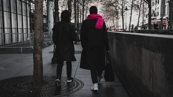 Scatto selettivo di due persone che indossano cappotti neri che camminano vicino ad alberi ed edifici — Foto Stock