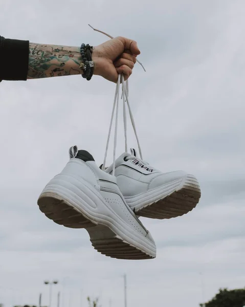 Vertical closeup shot of a male with tattoos holding a pair of white sneakers — Stock Photo, Image