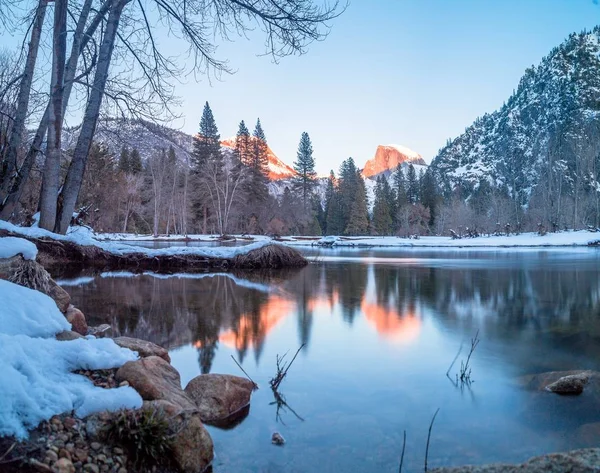 A lake surrounded by rocks, trees and mountains over Half Dome in Yosemite during winter — 스톡 사진