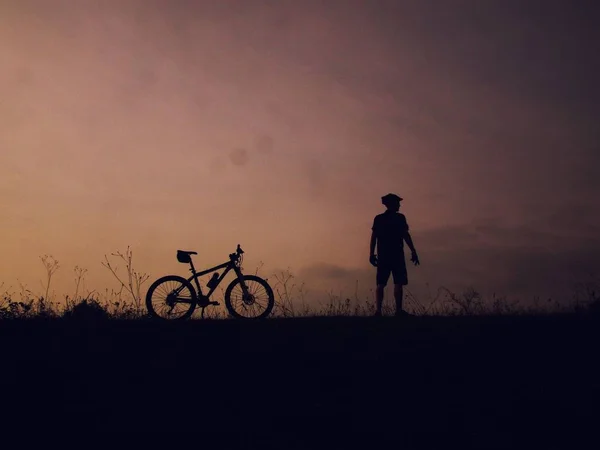 Silueta de un macho parado cerca de una bicicleta de montaña en una colina con un cielo púrpura en el fondo — Foto de Stock