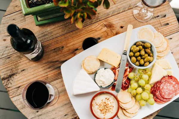 Wide shot of fruits and meat on white ceramic plate beside wine glasses and bottle on a wooden — Stock Photo, Image