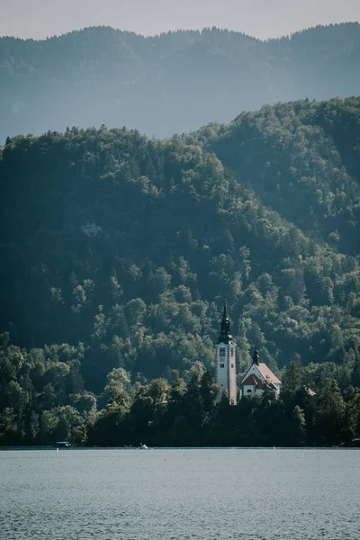 A vertical shot of sea with a house surrounded by trees in the distance near forested mountains