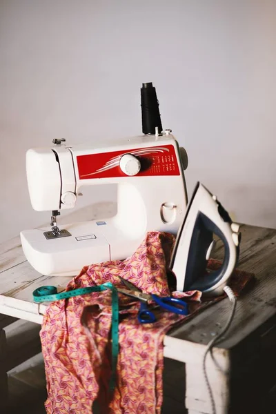 Primer plano de una máquina de coser blanca, textil, tijeras, cinta métrica y plancha en una mesa —  Fotos de Stock