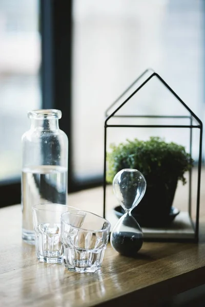 Gros plan sélectif d'un sablier, pot de fleurs dans un verre en forme de maison et verres à boire — Photo
