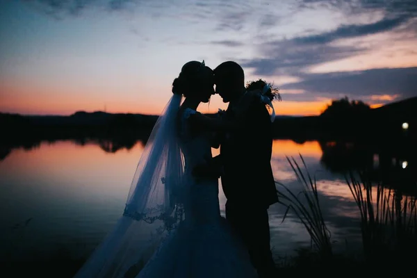 Silhouettes du marié et de la mariée se tenant passionnément près d'un lac au coucher du soleil — Photo