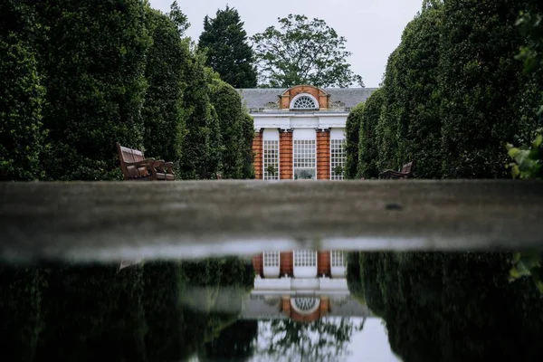 Casa marrón y blanca rodeada de árboles y bancos en un jardín con sus reflejos —  Fotos de Stock