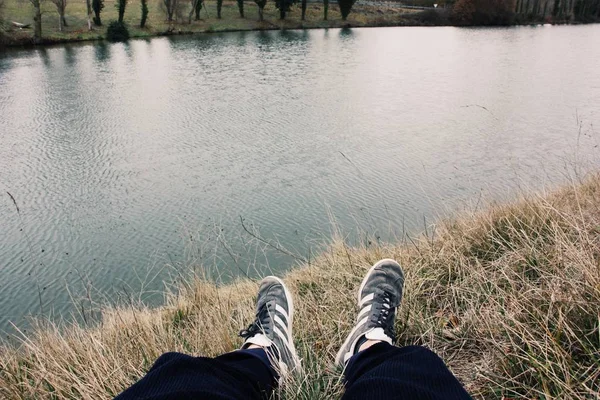 Leicht Nahaufnahme einer Person in Turnschuhen, die auf dem Gras am See sitzt — Stockfoto
