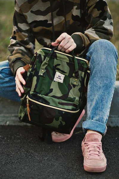 Osoba v vojenské mikině, modrých džínách a růžových teniskách držící batoh — Stock fotografie