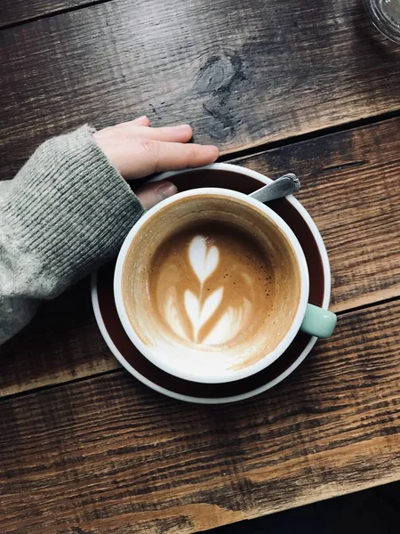 Overhead vertical shot of a person's hand near the latte art coffee on a wooden surface — Stock Photo, Image