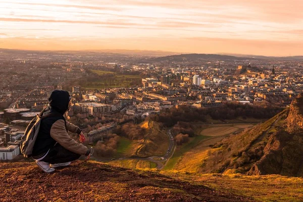 A person sitting at the top of a mountain surrounded by buildings in a city during sunset — Stock Photo, Image