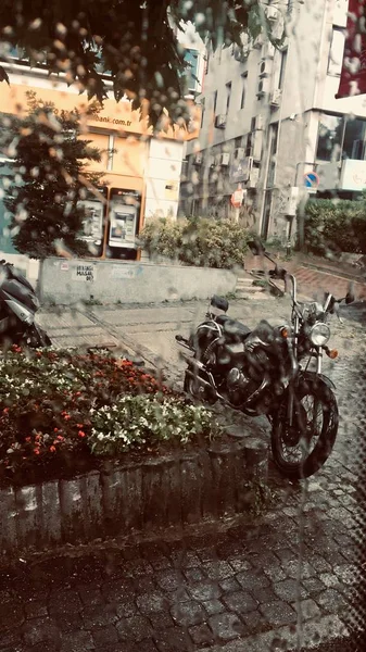 Vertical shot of a black motorcycle parked on a pavement near buildings during rain — Stock Photo, Image