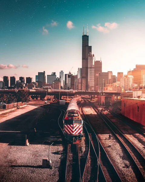 Tiro vertical de un tren sobre raíles cerca de rascacielos bajo un cielo azul al atardecer en una ciudad —  Fotos de Stock