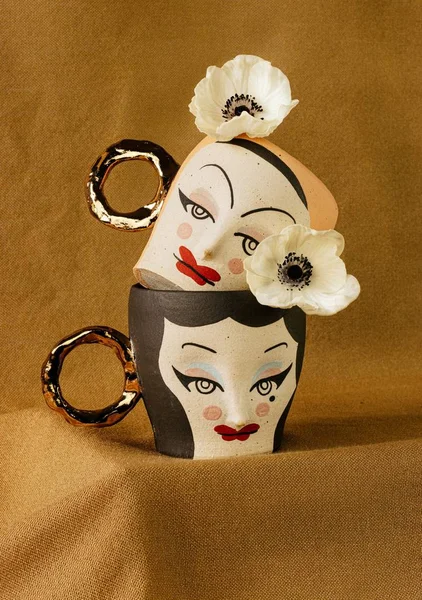 Vertical shot of black and peach mugs with female faces with white flowers on a brown background — Stock Photo, Image