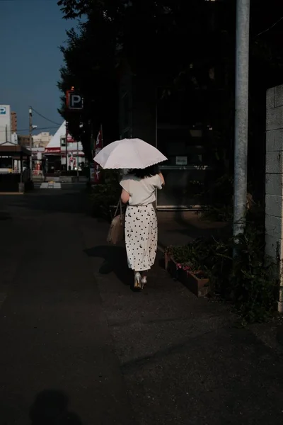 Κάθετη βολή θηλυκό φορώντας ένα πουκάμισο και μια φούστα κρατώντας ομπρέλα περπατώντας κατά μήκος ενός δρόμου με δέντρα — Φωτογραφία Αρχείου