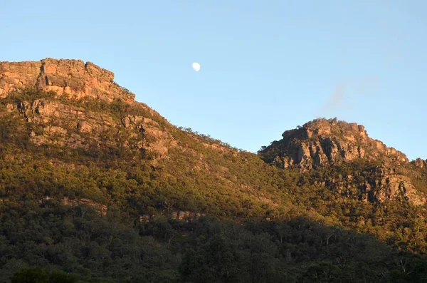 A beautiful shot of forested mountains under a clear sky with a visible moon at daytime