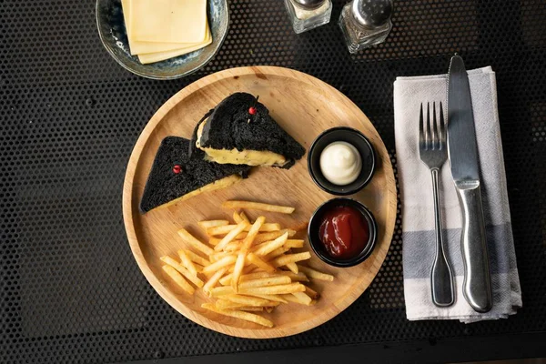 Overhead shot of black toasts with cheese near french fries and sauces on a wooden plate