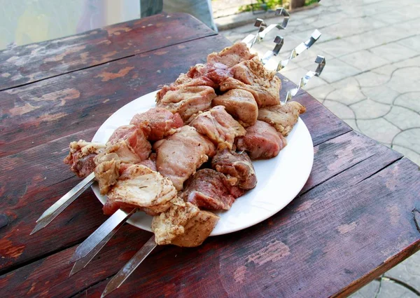 Placa blanca llena de brochetas y carne sazonada lista para asar en una vieja mesa de madera —  Fotos de Stock