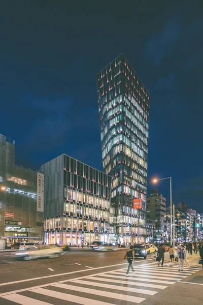 A vertical shot of tower blocks in a city full of lights and people walking in the street at night