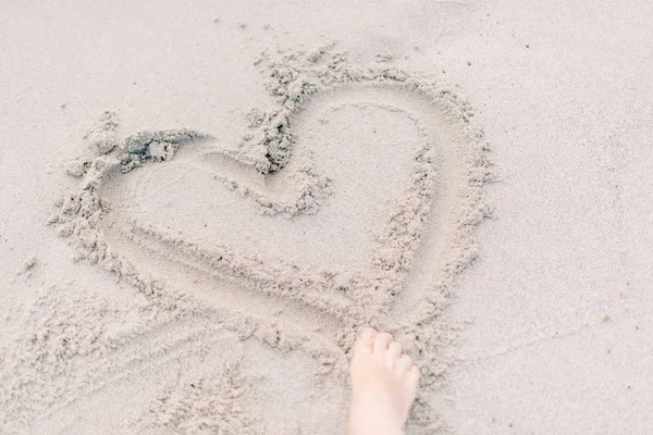 A close shot of a person drawing a heart shape on a sandy surface