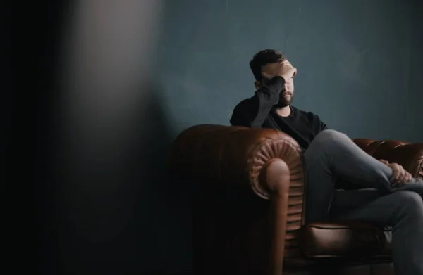 A selective shot of a depressed male sitting on a brown couch against a gray wall